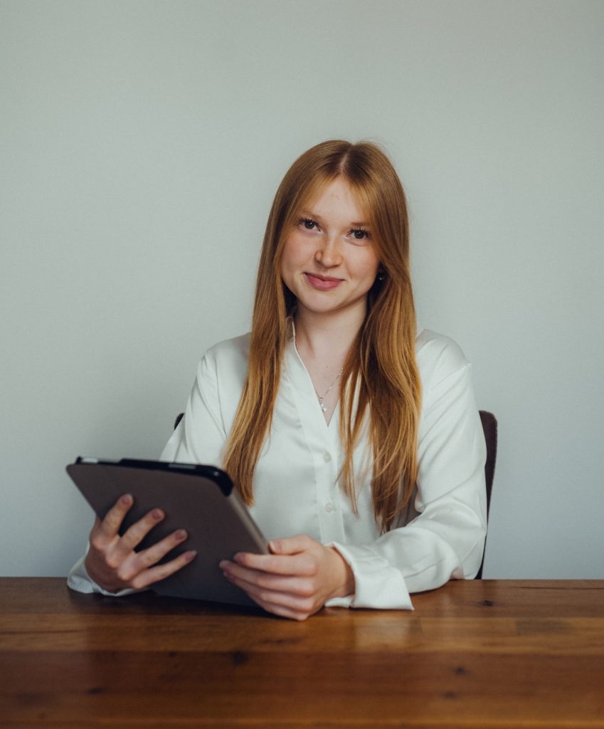 Junge Frau mit langen roten Haaren in weißer Bluse hält Tablet am Holztisch